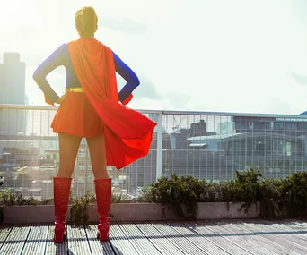 A woman in a superhero costume stands on a rooftop, gazing at the city skyline, with a red cape flowing behind her.