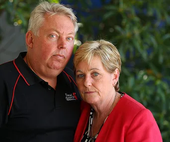 A concerned man and woman standing close, looking towards the camera, with greenery in the background.