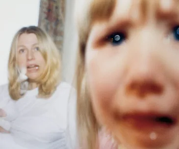 Blurry image of a distressed child close-up and a concerned woman in the background, both in a domestic setting.