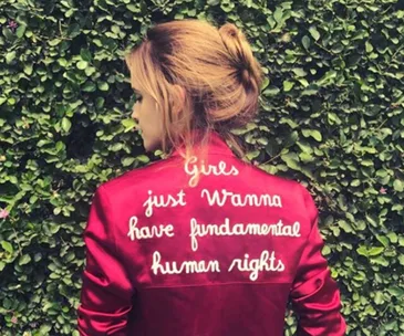 Red jacket with “Girls just wanna have fundamental human rights” text, worn by woman, standing against greenery.