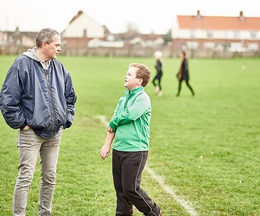 Man and boy talking on a grassy field with others in the background.