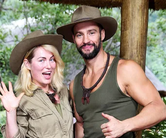 Two people smiling, wearing hats, outdoors with greenery in the background.
