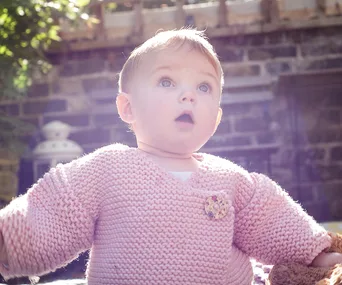 A baby in a pink knitted sweater with a surprised expression, sitting outside with a stone wall in the background.
