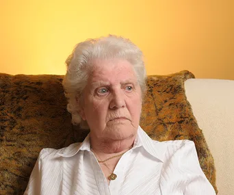 Elderly woman with white hair sitting, wearing a white blouse, looking to the side against an orange backdrop.