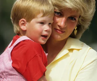 Mother in yellow cuddling a child in red overalls, outdoors.