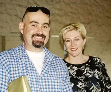 Man and woman smiling together, man in checkered shirt holding a golden package, indoors.