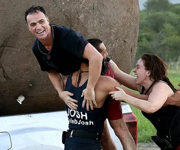 Group of people joyfully participating in an outdoor team challenge, assisting each other near a large boulder.
