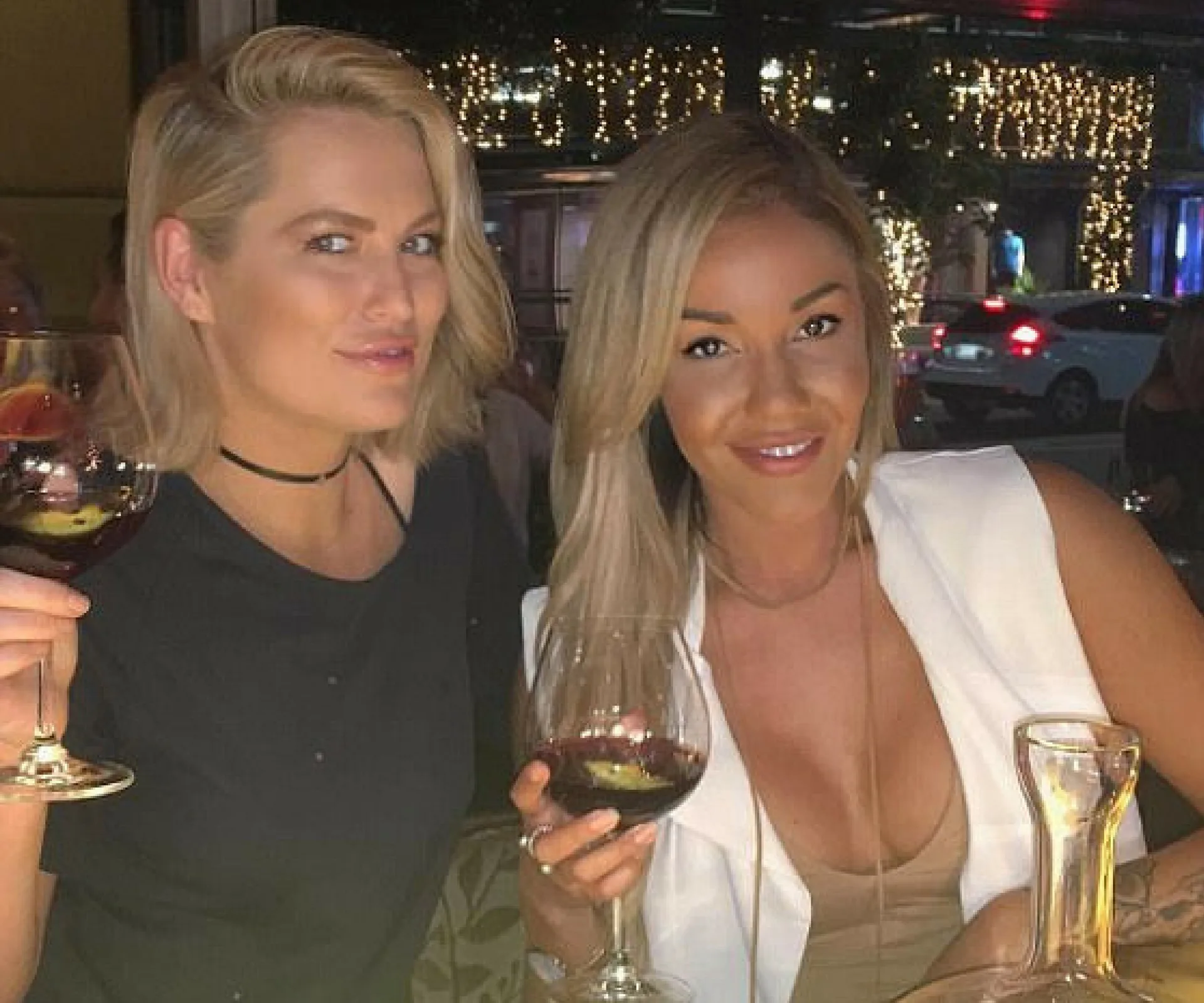 Two women smiling and holding glasses of red wine at an outdoor evening gathering.