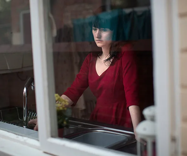 A woman in a red dress looks pensively through a window, standing near a kitchen sink.