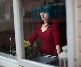 A woman in a red dress looks pensively through a window, standing near a kitchen sink.