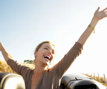 Person joyfully raises arms, smiling widely outdoors under a clear blue sky.