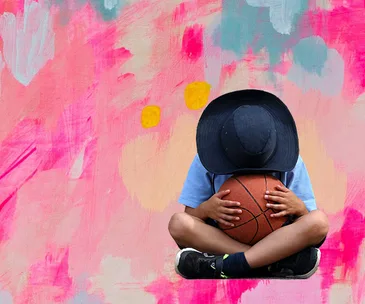 Child with wide-brim hat holding basketball, sitting against a colorful abstract background.