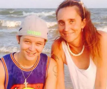 Two people smiling at the beach, one wearing a cap and jersey, the other in a tank top with long hair.