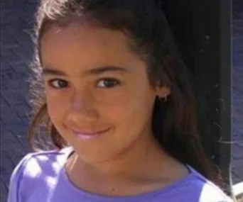 A young girl with long hair, wearing a purple shirt, smiling at the camera.