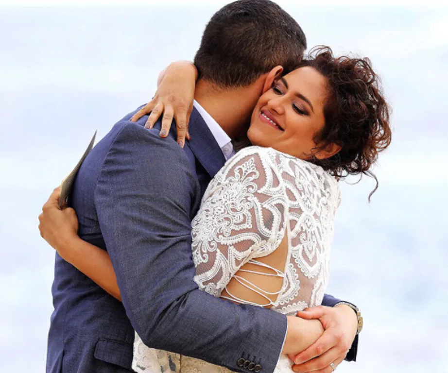 A couple in a warm embrace, the woman in a lace dress, smiles with her eyes closed against a serene backdrop.