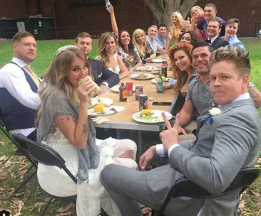 Group of people at an outdoor event seated around a table with food and drinks, some smiling and posing for a photo.