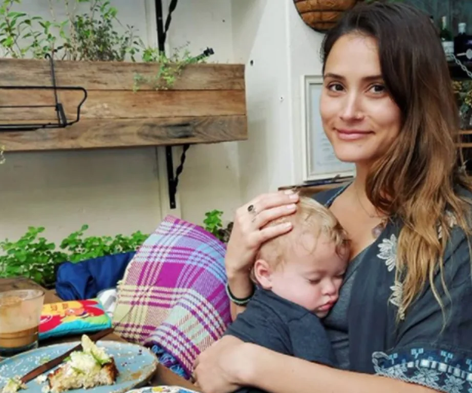 A woman sitting with a sleeping baby on her lap at an outdoor café, with food on the table in front of them.