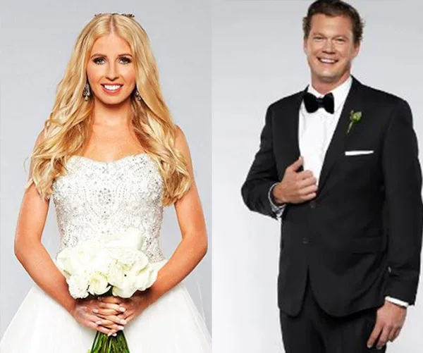 A woman in a wedding dress holds flowers beside a man in a black tuxedo, both smiling against a gray background.