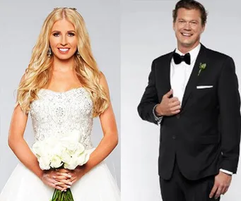 A woman in a wedding dress holds flowers beside a man in a black tuxedo, both smiling against a gray background.