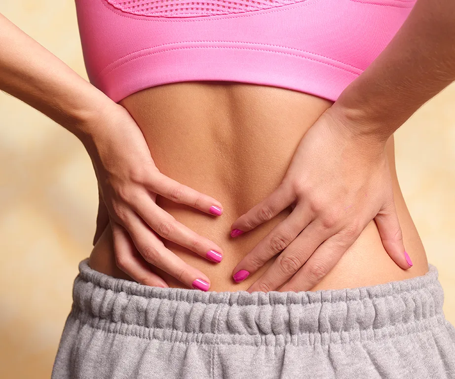 Person in pink top and grey pants holding lower back, suggesting back pain.