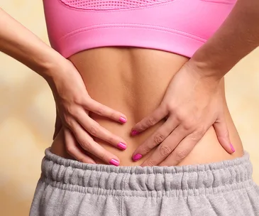 Person in pink top and grey pants holding lower back, suggesting back pain.