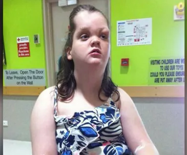 Teenage girl in hospital gown sitting in a medical room with green walls and informational signs.