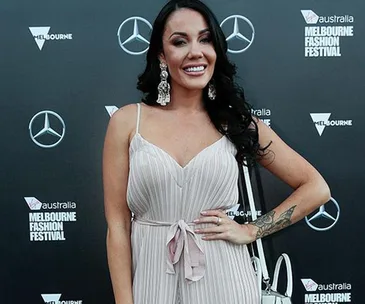 Woman in light dress posing at Melbourne Fashion Festival with branded backdrop.
