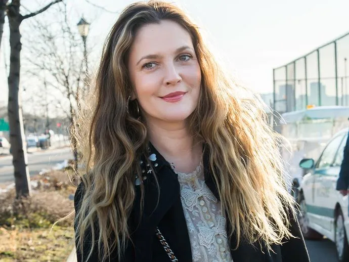 A person with long wavy hair and a lace top smiles outdoors on a sunny day.
