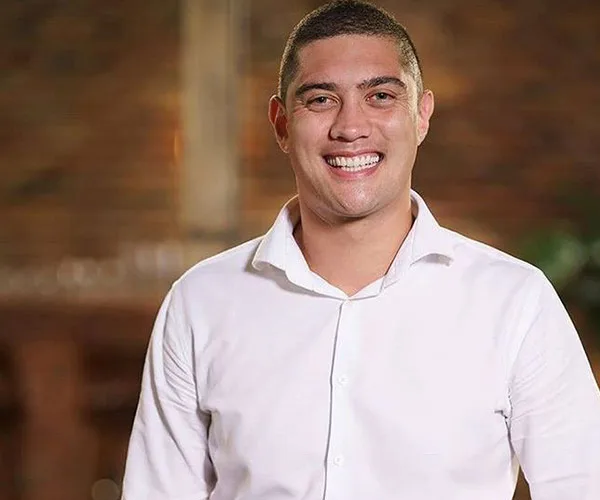 Man in a white shirt smiling against a blurred brick background.
