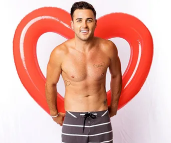 Shirtless man with tattoos stands in front of a red heart-shaped inflatable, wearing striped shorts and smiling at the camera.