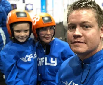 People in blue iFLY suits and orange helmets, smiling indoors, with a man in the foreground.