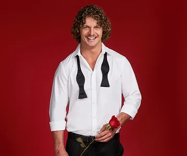 Man in a white shirt with a black bow tie undone, holding a red rose, against a red background.