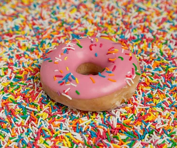 A pink frosted doughnut with colorful sprinkles sits on a bed of mixed sprinkles.