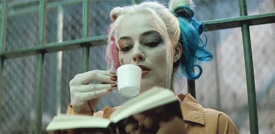 A person with colorful hair sips coffee while reading a book, sitting in what appears to be a confined space.