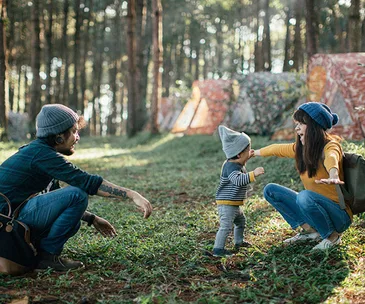 Family with toddler outdoors in a forest, surrounded by trees and tents, all wearing beanies and smiling.