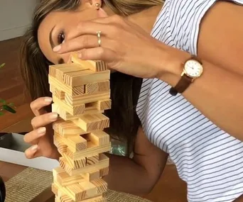 A person playing Jenga, wearing a striped shirt and a watch, with an engagement ring visible on their finger.