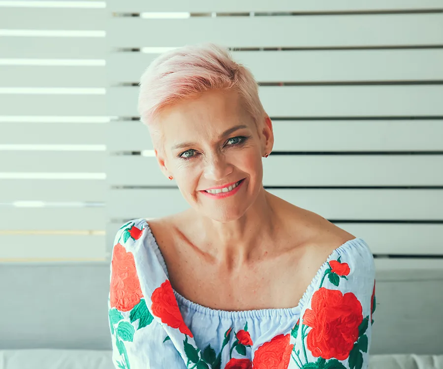 A woman with short, pink hair smiling, wearing an off-shoulder floral dress, against a slatted white background.