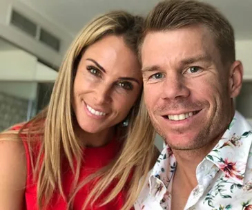 A smiling couple takes a selfie in a bright room, with a woman in a red top and a man in a floral shirt.