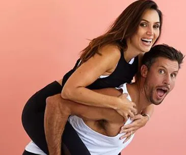 Couple playfully posing; woman piggybacks on man, both smiling against a peach-colored background.