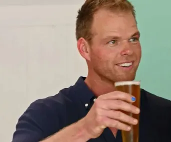 A man in a navy shirt holding a beer, with blue nail polish on one finger, smiling at something off-camera.