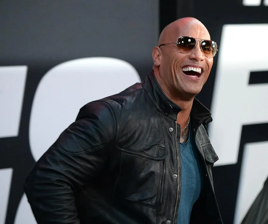 Dwayne "The Rock" Johnson wearing sunglasses and a leather jacket, smiling, at a public event.