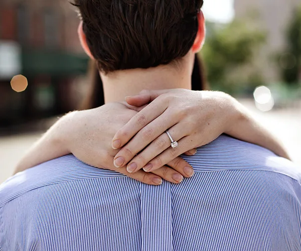 Close-up of a couple hugging; focus on a woman's hands with an engagement ring on a man's shoulder, outdoors.