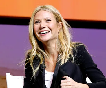 A person smiling and sitting on stage during a talk event, with a vibrant orange and purple background.