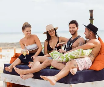 "Four people relaxing on a beach lounge, smiling and chatting, with drinks and pillows, under a bright sky."