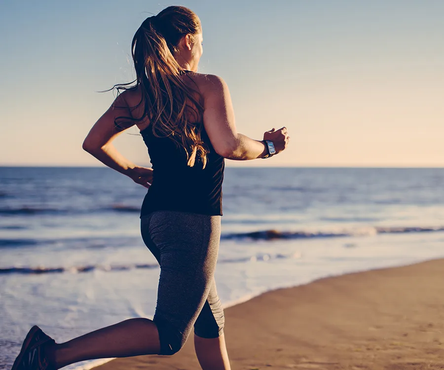 Woman jogging on beach at sunrise, wearing fitness tracker.