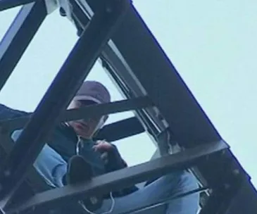 A person sitting on a metal structure, viewed from below, wearing a cap, jacket, jeans, and holding an object.