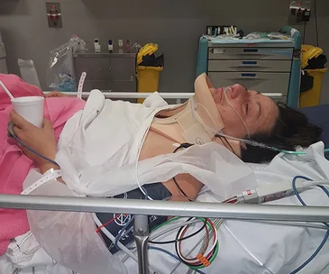 Patient in a hospital bed with a neck brace and medical equipment, holding a cup, surrounded by medical supplies.