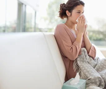A woman sitting on a couch, holding a tissue to her nose, wrapped in a blanket, with a box of tissues nearby.