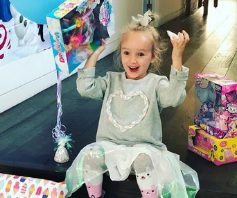 Young girl excitedly holding a doll box, sitting with birthday presents around her, including a balloon and colorful wrapping.
