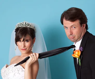 Bride pulling groom's tie with playful expression, both in wedding attire against blue background.